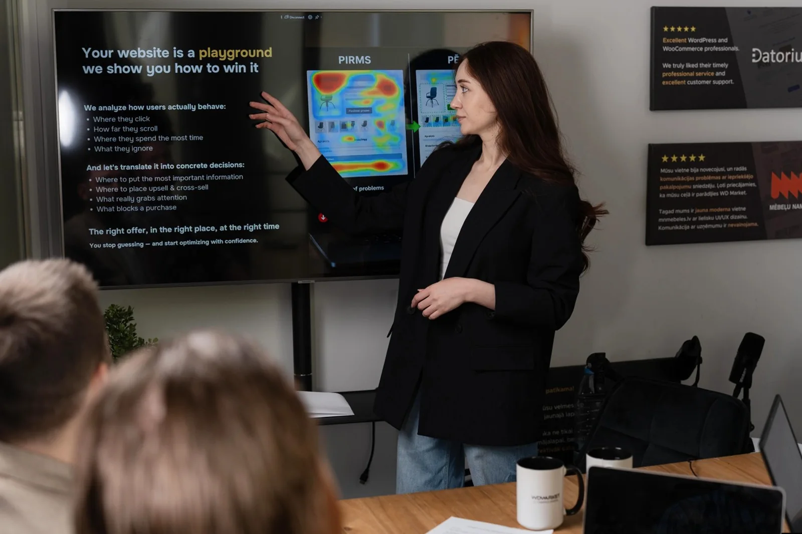 Instructor presenting to a small group, pointing at a screen that compares before and after click heatmaps of an online store page