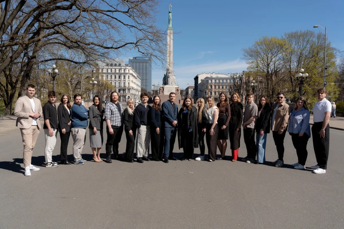 The eComCademy team standing in a long line outdoors in Riga, with the Freedom Monument behind them on a clear spring day
